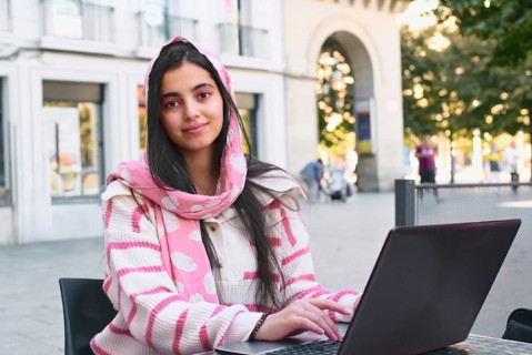 A arab student woman wearing a pink scarf and striped sweater is seated at an outdoor cafe, working