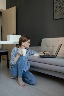 A woman sitting on the floor by a sofa, working on a laptop. She holds a coffee mug and listens