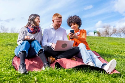 Three university students working together on a project outdoors, using a laptop, tablet and phone,