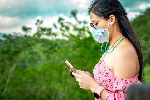 Latin woman with mask checking cell phone outdoors. Cute girl in glasses checking her cell phone