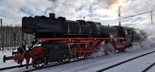A black, historic steam locomotive emits smoke and stands on snow-covered tracks in winter,
