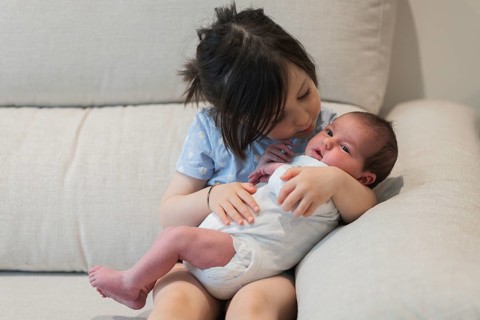 A touching moment as a big sister lovingly embraces her newborn sibling on a sofa. The young
