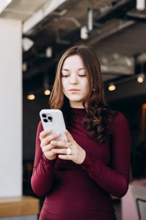 A young female student with long hair smiles as she looks at her smartphone, wearing a maroon dress