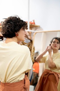 A woman stands in front of a large mirror, adjusting her earrings and admiring her reflection. The