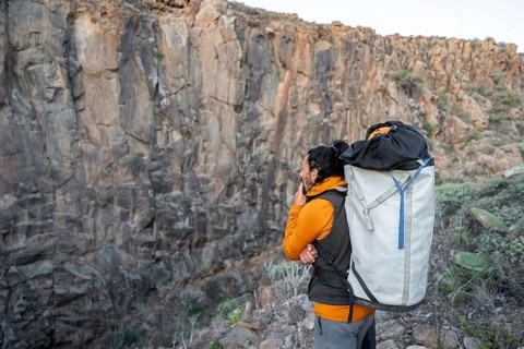 A climber with an orange jacket and a large backpack pauses to observe a towering rock face,