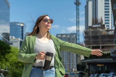Businesswoman hailing a taxi in a modern city center, holding a coffee thermos and wearing