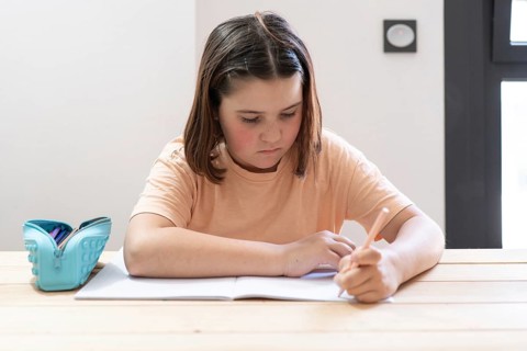 A young girl with brown hair concentrates on writing in her notebook. She sits at a wooden table