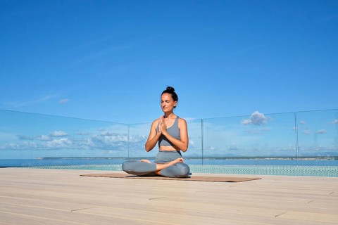 A woman practices meditation on a rooftop deck, seated in a lotus position. The clear blue sky and