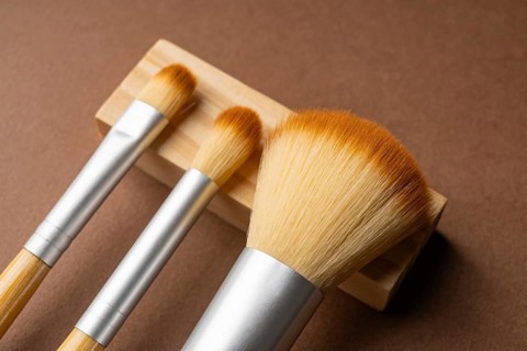 From above, three bamboo makeup brushes with silver metal accents rest on a light wood holder