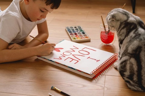 A young boy, lying on the wooden floor, draws a heartfelt Mother's Day card with watercolors. A