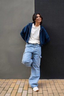 A person leans against a two-tone wall, wearing a casual outfit with a blue hoodie, white T-shirt,