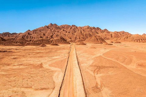 A solitary dirt road stretches through a vast, arid desert landscape leading towards striking,