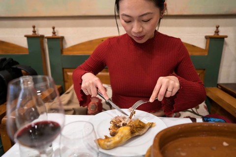 An Asian female tourist in Segovia, Spain, enjoys a traditional roast suckling pig dish. Seated in