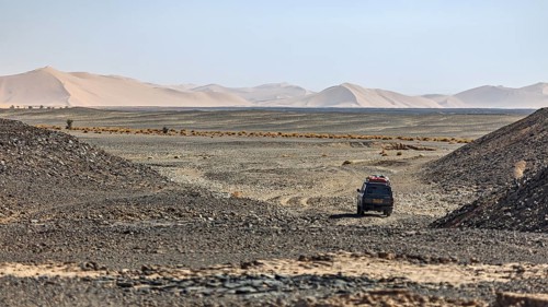 A vehicle moves through a barren desert landscape behind sand dunes, With off-road vehicle through