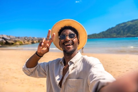 Young man wearing hat and sunglasses gesturing victory sign while taking selfie on a tropical beach