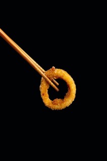 A golden, crispy onion ring delicately held between wooden chopsticks, set against a dark