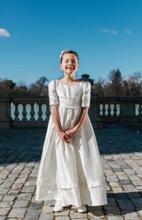 A young girl stands outdoors in a beautiful First Communion dress, radiating joy on a sunny day.
