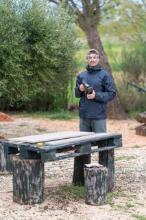 A man stands outdoors near a wooded area, holding a jigsaw for a carpentry project. He sports