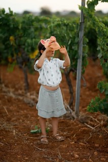 A young child stands in a vineyard, playfully holding a pig mask over their head. The scene