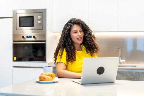 Female latin young freelancer having breakfast while attending online meeting from home sitting in