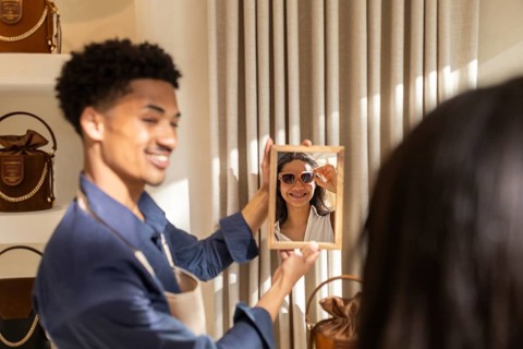 A cheerful customer in a stylish bag store tries on sunglasses while an employee holds a mirror,