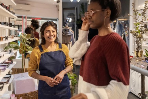 A smiling store assistant in a modern sunglasses shop assists a customer trying on glasses. The