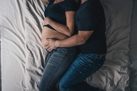 A couple is lying in bed, with the man embracing the pregnant woman and placing his hand on the