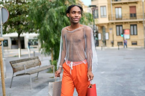 Confident man in a stylish mesh top and vibrant pants holds shopping bags while strolling in an