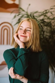 A teenager with blonde hair smiles thoughtfully, touching her face. She's wearing a green top in a