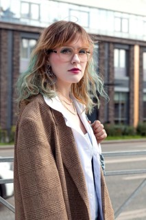 Stylish woman with wavy hair, wearing glasses and a blazer, poses confidently in an urban