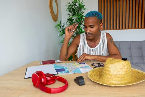 An LGBTQIA+ man with vibrant blue hair is preparing for a trip. He is seated at a table with a map,