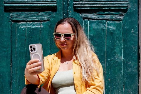 A smiling woman tourist wearing sunglasses and a yellow shirt takes a selfie on a colorful street,
