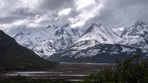 Snow-covered mountain chain of the Andes with pointed peaks under cloudy skies, wooded slopes and