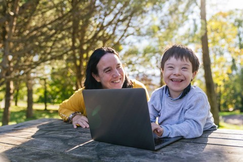 A smiling boy with Down syndrome and his mother enjoying a learning session outdoors with a laptop.