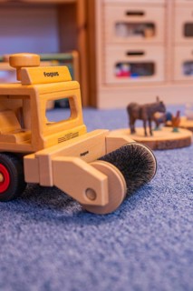 A wooden toy sweeper stands on blue carpet, with small animal figures in the background, infant and