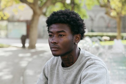 Portrait of a young black man seated outdoors in a park, looking away with a thoughtful expression.