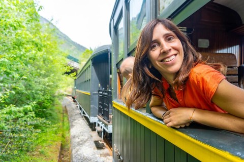 A woman is smiling as she looks out the window of a train. The train is surrounded by trees and he