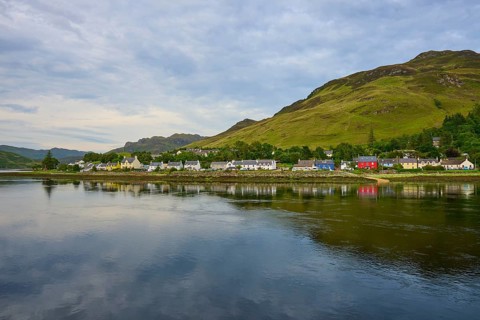 A calm lake with mountains and houses on the shore surrounded by green trees under a cloudy sky,