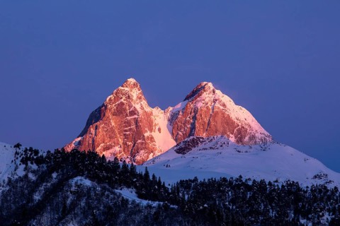 Snow-capped peaks glow with warm sunset hues against a vibrant blue sky in Georgia A dense forest