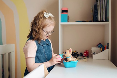 A young girl with Down syndrome happily plays with toys in a cozy room. She is engaged and smiling,