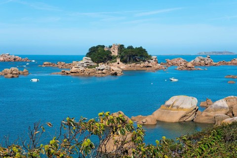 Rocky island with trees and old building, surrounded by clear blue sea under a sunny sky, granite