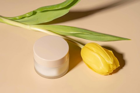 A minimalist scene with a skincare cream jar next to a yellow tulip on a beige background. The