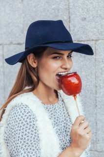 A smiling woman in a fashionable hat takes a bite of a glossy red candied apple. The image captures