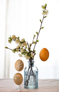 Blown out Easter eggs hang from blossoming branches that stand in a light blue vase against a light