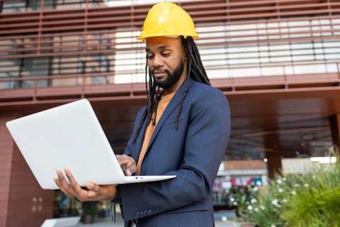 Cuban businessman wearing a yellow hard hat and navy suit, engaged with a laptop outside a modern