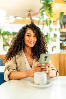 Vertical portrait of a chic beauty latin woman using phone sitting on a cafeteria drinking hot