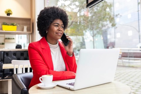 Confident woman in a vibrant red blazer talks on her phone while working on a laptop in a bright,