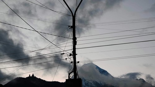 A power pole with cables in front of a snowy mountain, Kazbek with Gergeti church under dramatic