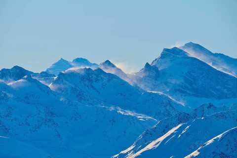 Snowy mountain peaks backlit with flying snow, against a clear blue sky in a wintry landscape,