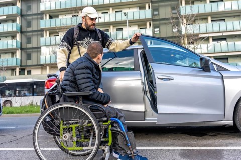 Caregiver assisting a young disabled man in a wheelchair while entering a car parked on a city